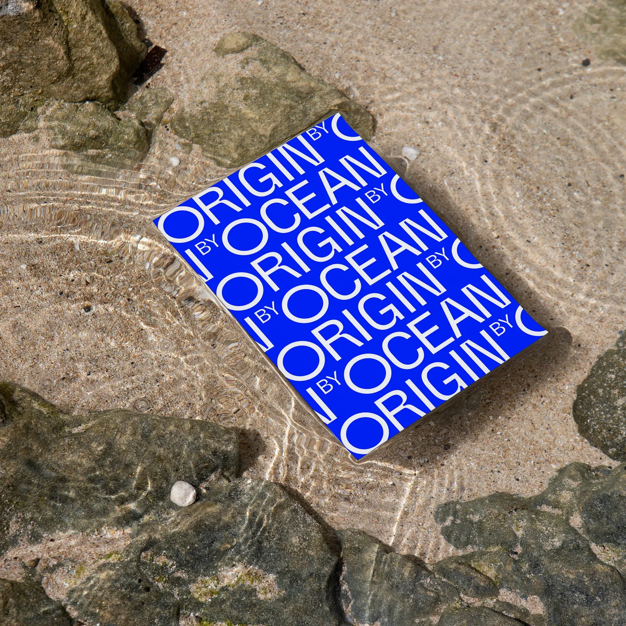 Origin by Oceanin brändikirja, jonka kannessa toistuu sininen logo, matalassa merivedessä kivillä.