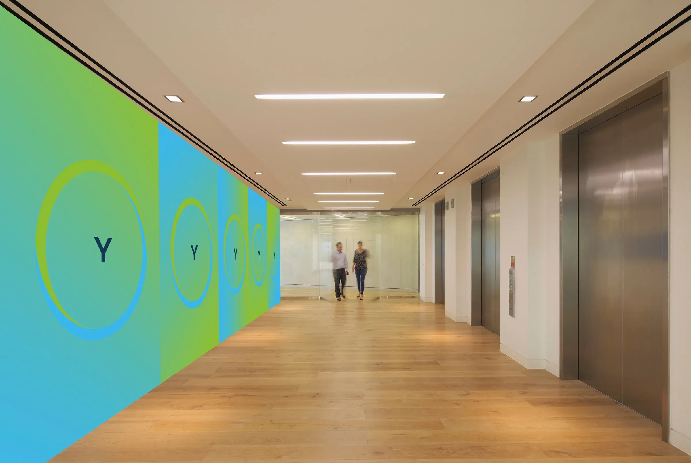 Office corridor with a wall of blue and green panels, each carrying the Y in a ring, and two people walking.