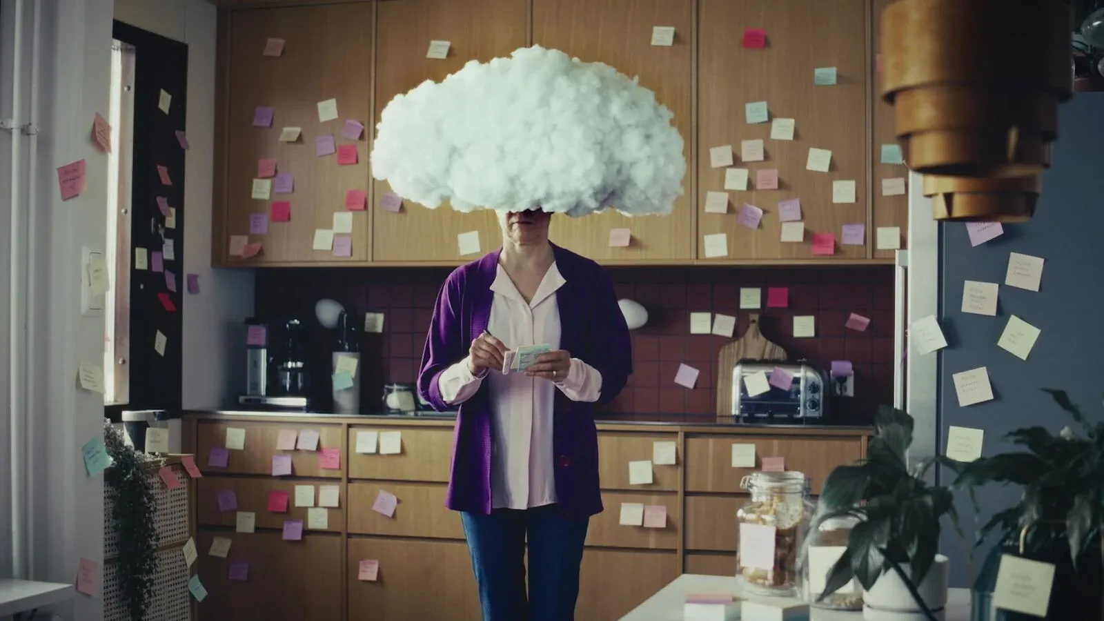 A woman in an office kitchen covered in sticky notes, her face hidden by a white cloud.