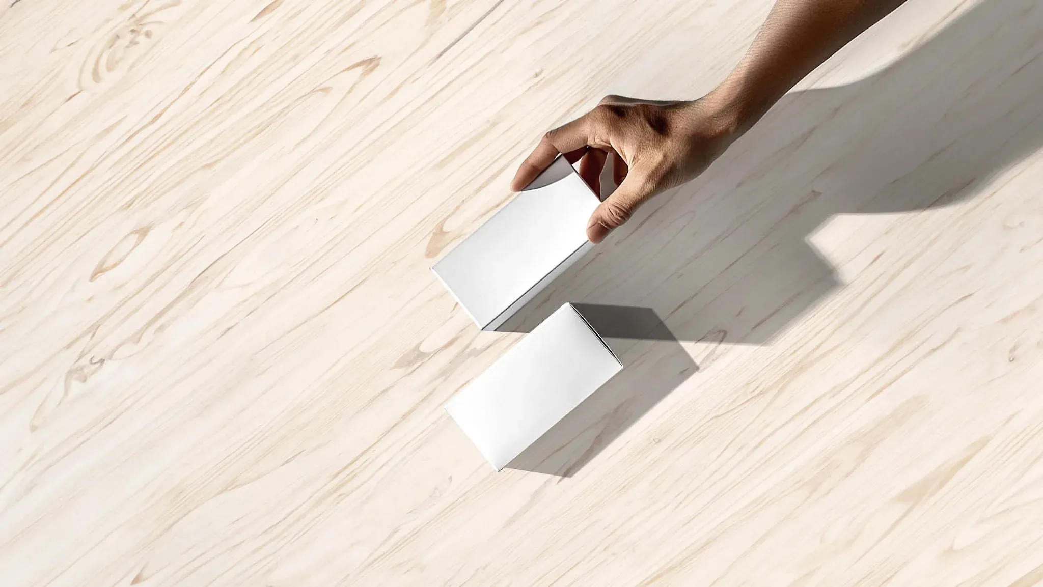 A hand placing a white box beside another on a pale wooden surface.